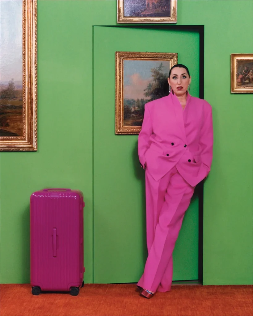 Rossy de Palma a magenta kufor RIMOWA pred zelenou stenou s obrazmi.