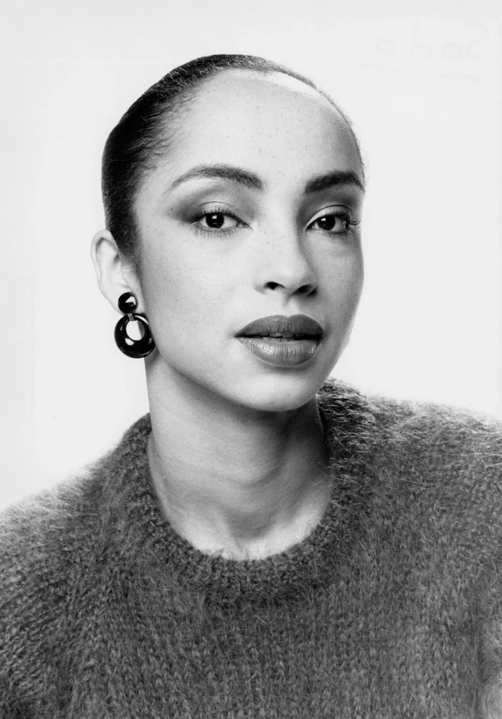 Čiernobiela portrétna fotografia Sade Adu v pletenom svetri s masívnymi náušnicami.