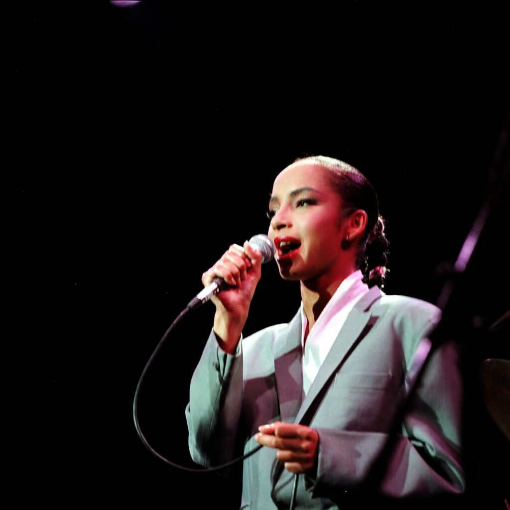 Sade Adu počas živého vystúpenia v elegantnom sivom saku s mikrofónom.