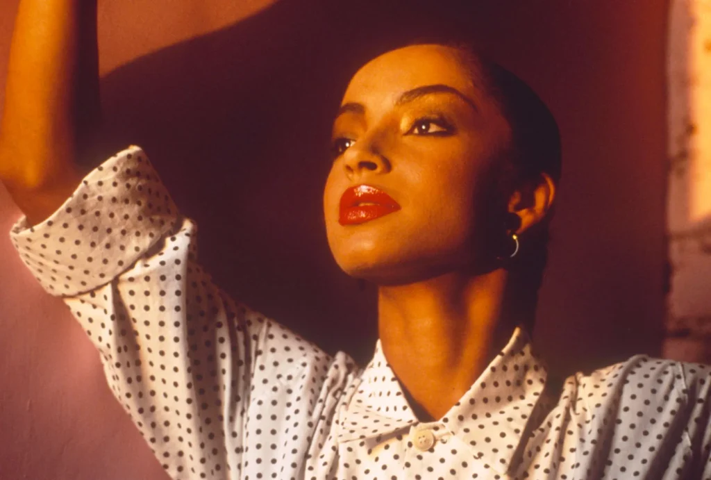Speváčka Sade Adu v ikonickej bodkovanej košeli s výrazným červeným rúžom.