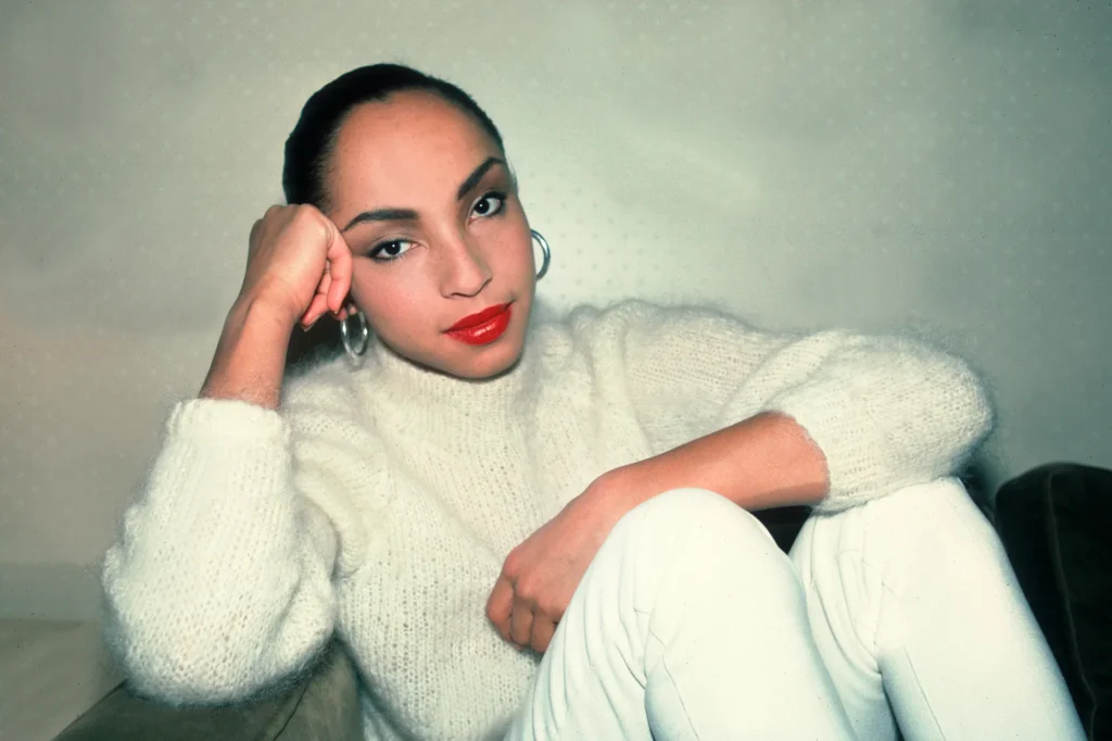 Minimalistický portrét Sade Adu v bielom pletenom svetri a strieborných kruhových náušniciach.