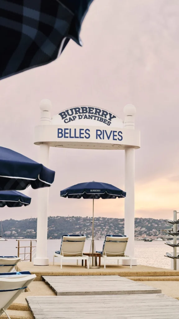 Plážové mólo s ikonickým nápisom Burberry Cap d'Antibes Belles Rives, tmavomodrými slnečníkmi a lehátkami počas západu slnka.