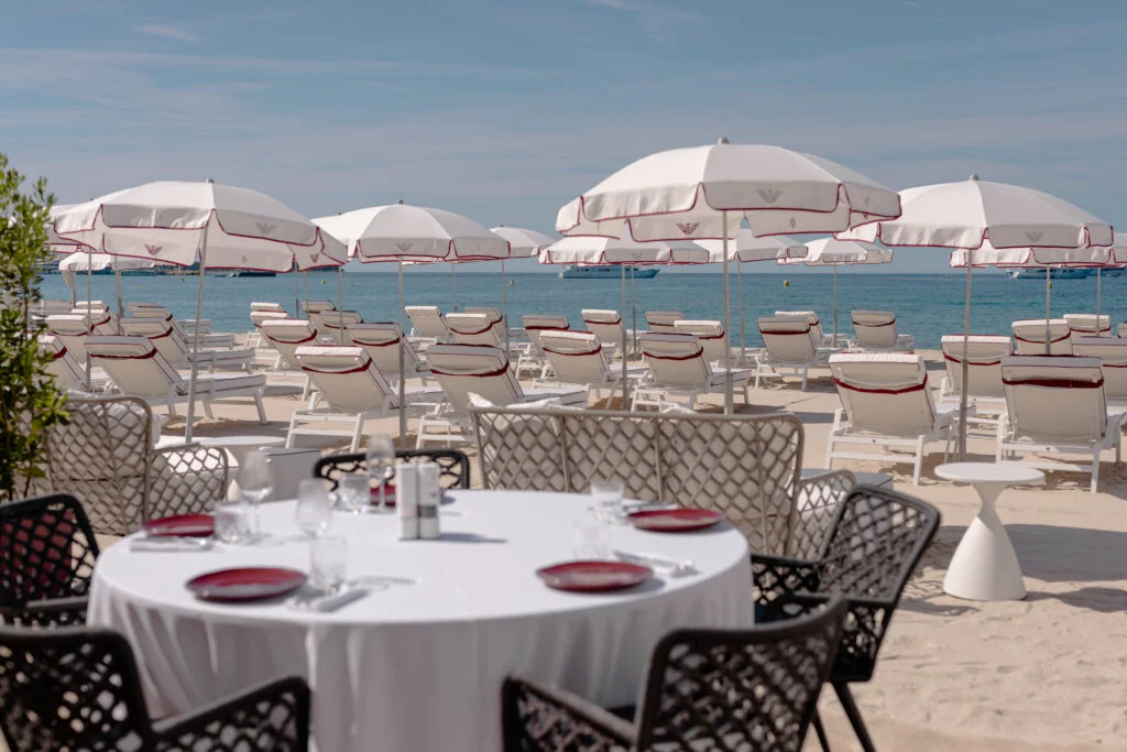 Luxusná piesočná pláž v Cannes s bielymi slnečníkmi a lehátkami s červeným lemovaním od značky Emporio Armani s prestretým stolom v popredí.