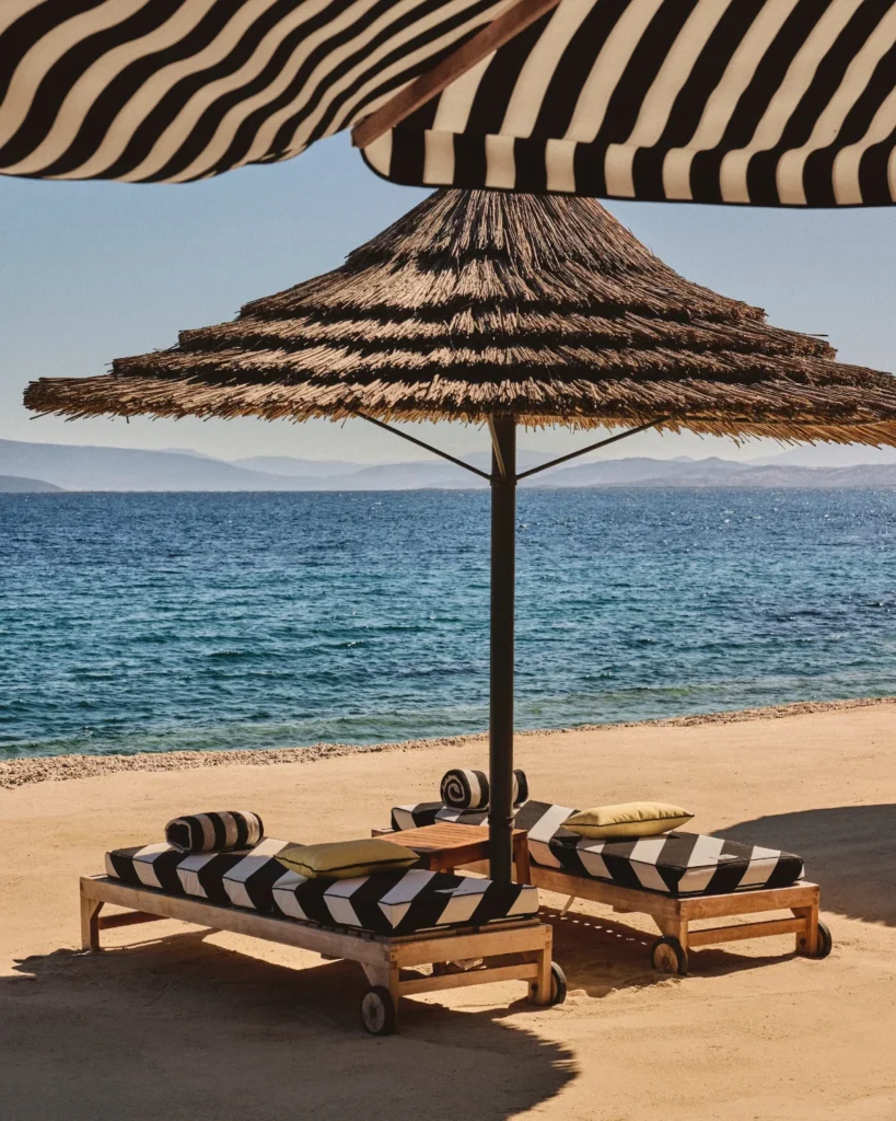 Slnečník zo slamy a plážové lehátka s čierno-bielym pásikavým vzorom od dizajnéra Jacquemus v rezorte Mandarin Oriental Bodrum pri mori.