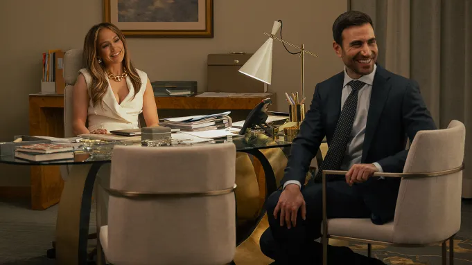 Usmievavá Jennifer Lopez sedí za pracovným stolom vo svojej kancelárii oproti Brettovi Goldsteinovi vo filme Office Romance (Pracovní romance).