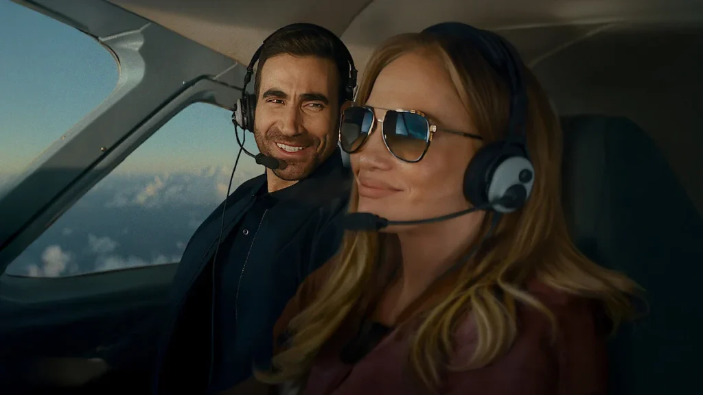Jennifer Lopez so slnečnými okuliarmi a Brett Goldstein s leteckými slúchadlami v kabíne lietadla vo filme Office Romance (Pracovní romance).