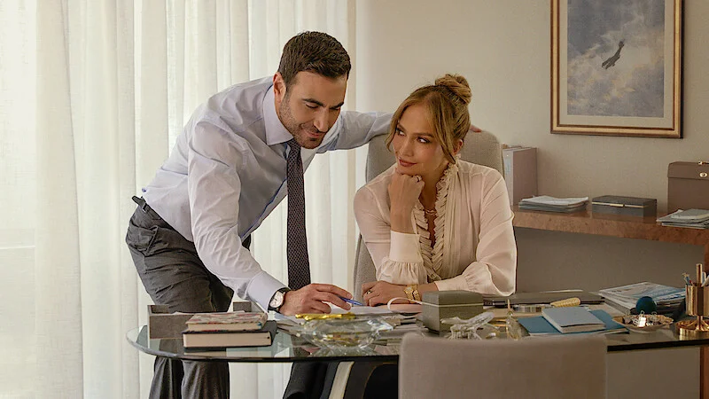 Brett Goldstein sa nakláňa nad pracovný stôl, za ktorým sedí Jennifer Lopez v scéne z filmu Office Romance (Pracovní romance).
