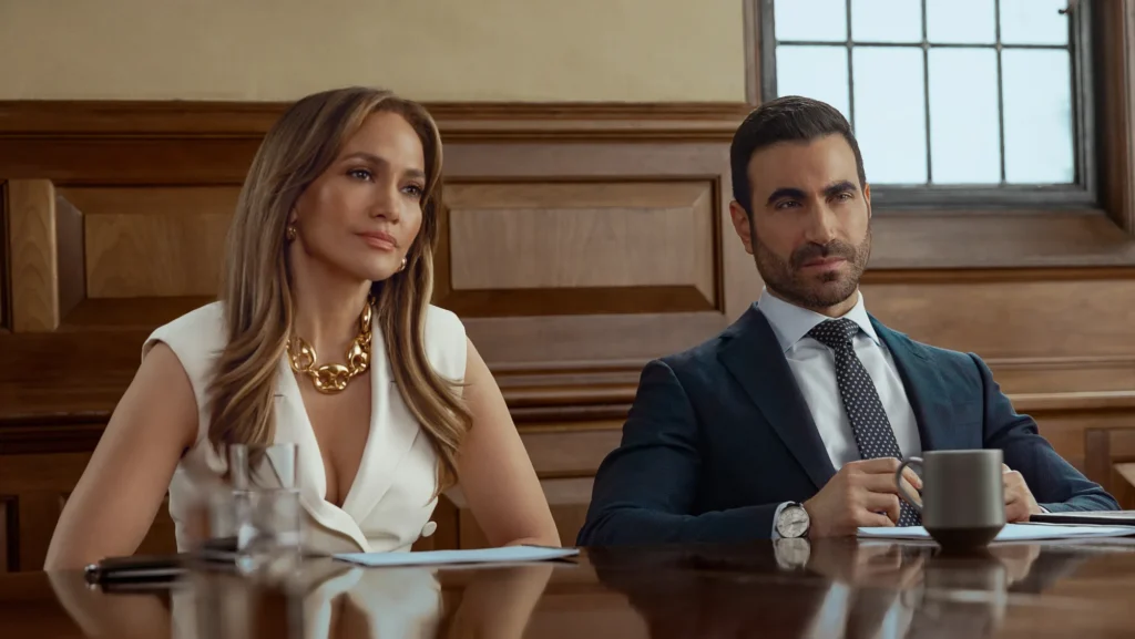 Jennifer Lopez a Brett Goldstein sedia vedľa seba za veľkým stolom v zasadacej miestnosti vo filme Office Romance (Pracovní romance).