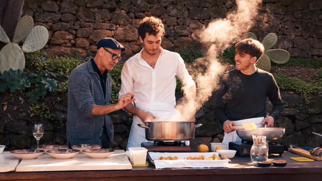 Stanley Tucci sleduje kulinársku prípravu jedla na otvorenom priestranstve s miestnymi obyvateľmi.